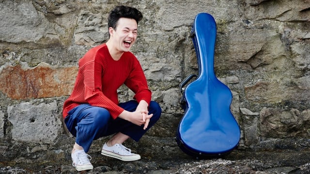 Sean Shibe laughing with guitar case in front of stone wall