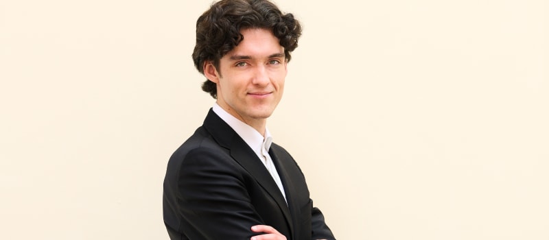 Ignas Maknickas (pianist) wearing a black suit standing, arms folder, on a beige background